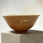Close-up of a brown ceramic bowl with gold kintsugi cracks, resting on a white display pedestal against a pale background.