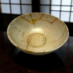 Close-up of a white ceramic bowl with gold repair lines (kintsugi) on a dark wooden surface.