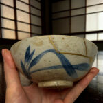 Close-up of a person holding a decorated ceramic bowl with a string tied around it, fingers visible at the edge.