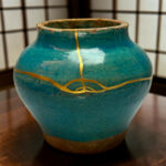 Close-up of a teal ceramic vase with gold crackle lines and a worn rim on a wooden surface behind it.