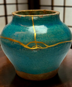 Close-up of a teal ceramic vase with gold crackle lines and a worn rim on a wooden surface behind it.