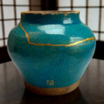 Close-up of a teal ceramic vase with gold decorative lines and a cracked glaze, sitting on a dark wooden surface. The background shows a blurred grid pattern.