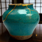 Close-up of a teal ceramic vase with gold crackle lines on a wooden table, blurred grid background behind it.