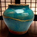 Close-up of a blue ceramic vase with a weathered brown rim and a yellow string wrapped around its midsection, background blurred grid pattern.