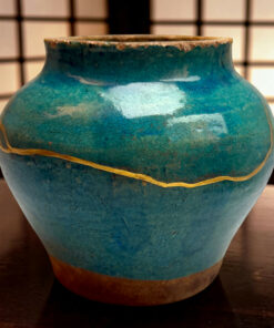 Close-up of a blue ceramic vase with a weathered brown rim and a yellow string wrapped around its midsection, background blurred grid pattern.
