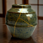Close-up of a green ceramic vase with a crack tied by a yellow cord, resting on a wooden surface138]