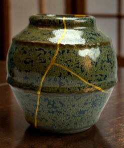 Close-up of a green ceramic vase with a crack tied by a yellow cord, resting on a wooden surface138]