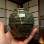 Close-up of a speckled green ceramic vase held in hands, with a gold cord wrapped around it. background shows wooden shelves.