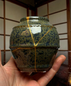 Close-up of a speckled green ceramic vase held in hands, with a gold cord wrapped around it. background shows wooden shelves.