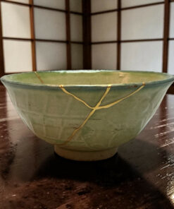 Close-up of a green ceramic bowl with gold repair seams on a dark wooden table, background a lattice screen
