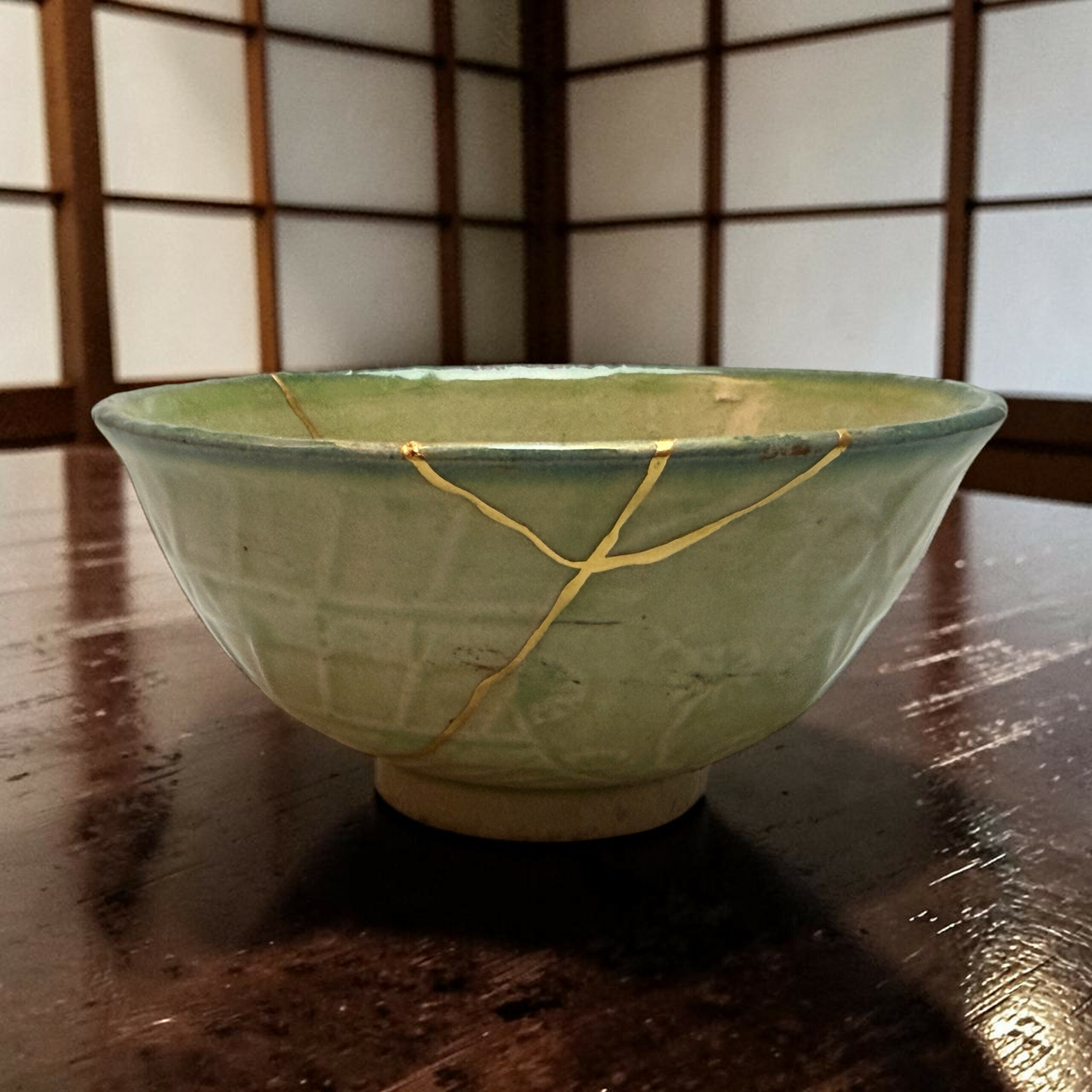 Close-up of a green ceramic bowl with gold repair seams on a dark wooden table, background a lattice screen