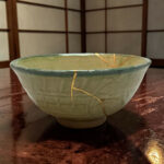 Close-up of a green ceramic bowl with a crack running across, resting on a dark wooden surface with a tan string laid across its rim.