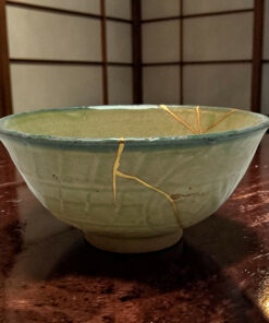 Close-up of a green ceramic bowl with a crack running across, resting on a dark wooden surface with a tan string laid across its rim.