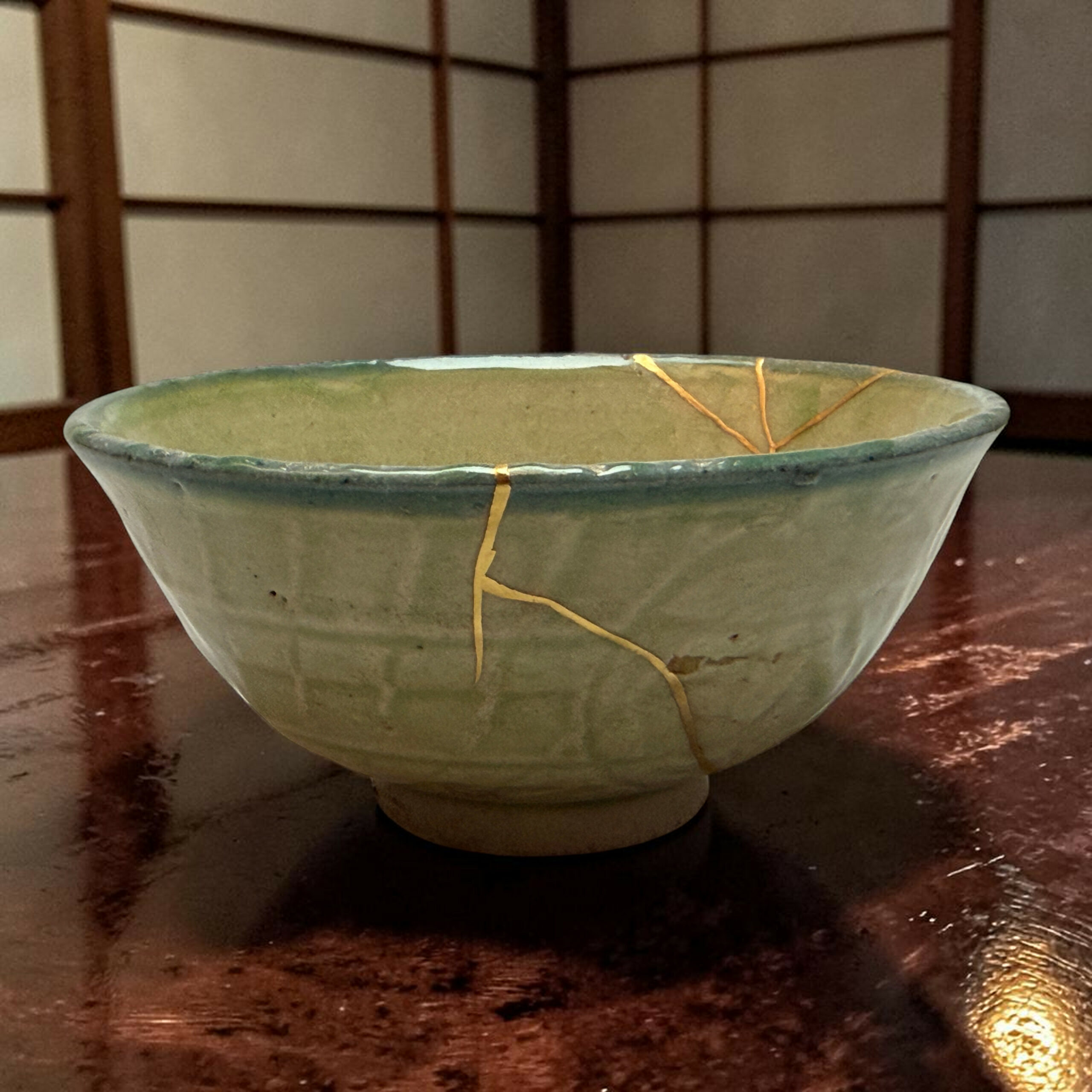 Close-up of a green ceramic bowl with a crack running across, resting on a dark wooden surface with a tan string laid across its rim.