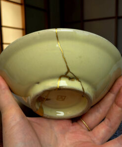 Close-up of a pale green ceramic egg repaired with gold seams (kintsugi), being gently held by hands.