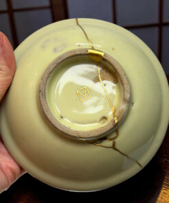 Light green ceramic plate with a long crack held by a hand; a gold repair seam runs along the crack earlier repaired in gold.