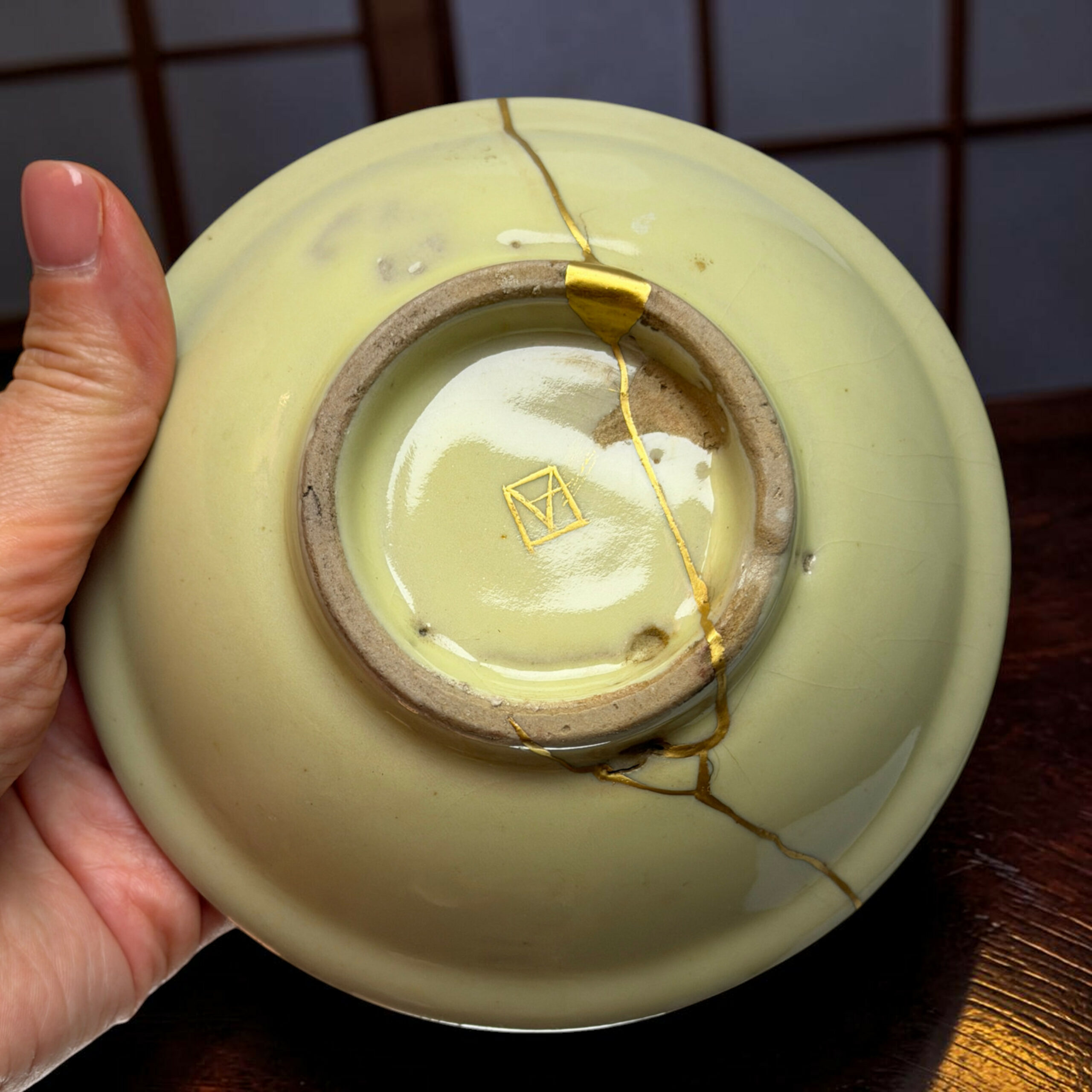 Light green ceramic plate with a long crack held by a hand; a gold repair seam runs along the crack earlier repaired in gold.