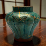 Close-up of a teal ceramic vase with gold kintsugi repairs and a wooden table surface in the background.