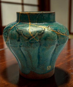 Close-up of a teal ceramic vase with gold kintsugi repairs and a wooden table surface in the background.