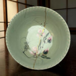 Close-up of a mint-green ceramic plate with a gold crack line and pink floral pattern on a wooden surface.