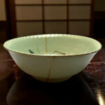White ceramic bowl with a visible hairline crack running from top to bottom, set on a dark wooden table with a blurred grid background behind it.
