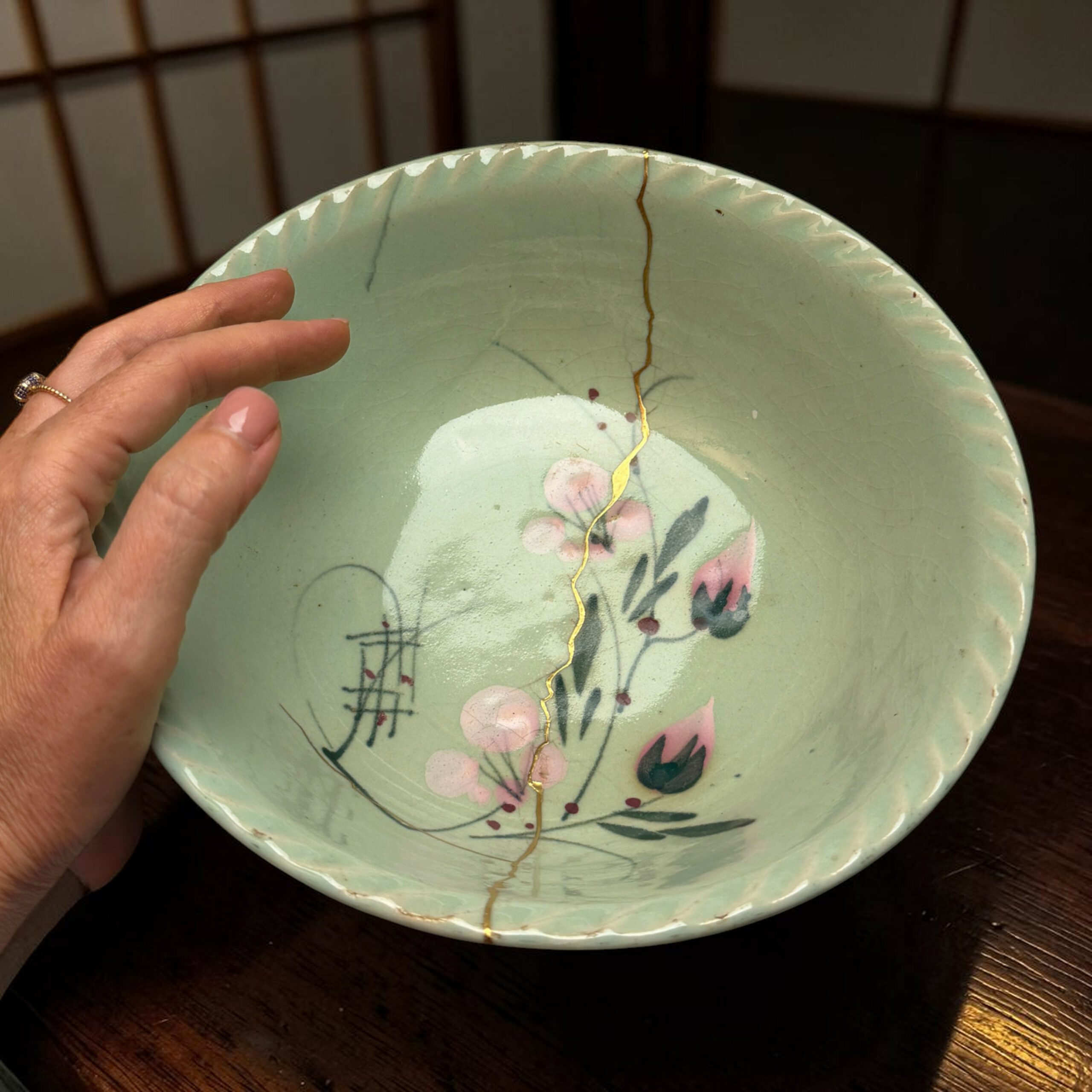 Hand resting near the edge of a pale green porcelain plate with pink floral design and a gold hairline crack, on a dark wooden table.
