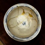 Close-up of a white ceramic plate with blue striped rim and gold repair seams (kintsugi) against a dark background, showing repaired cracks.",