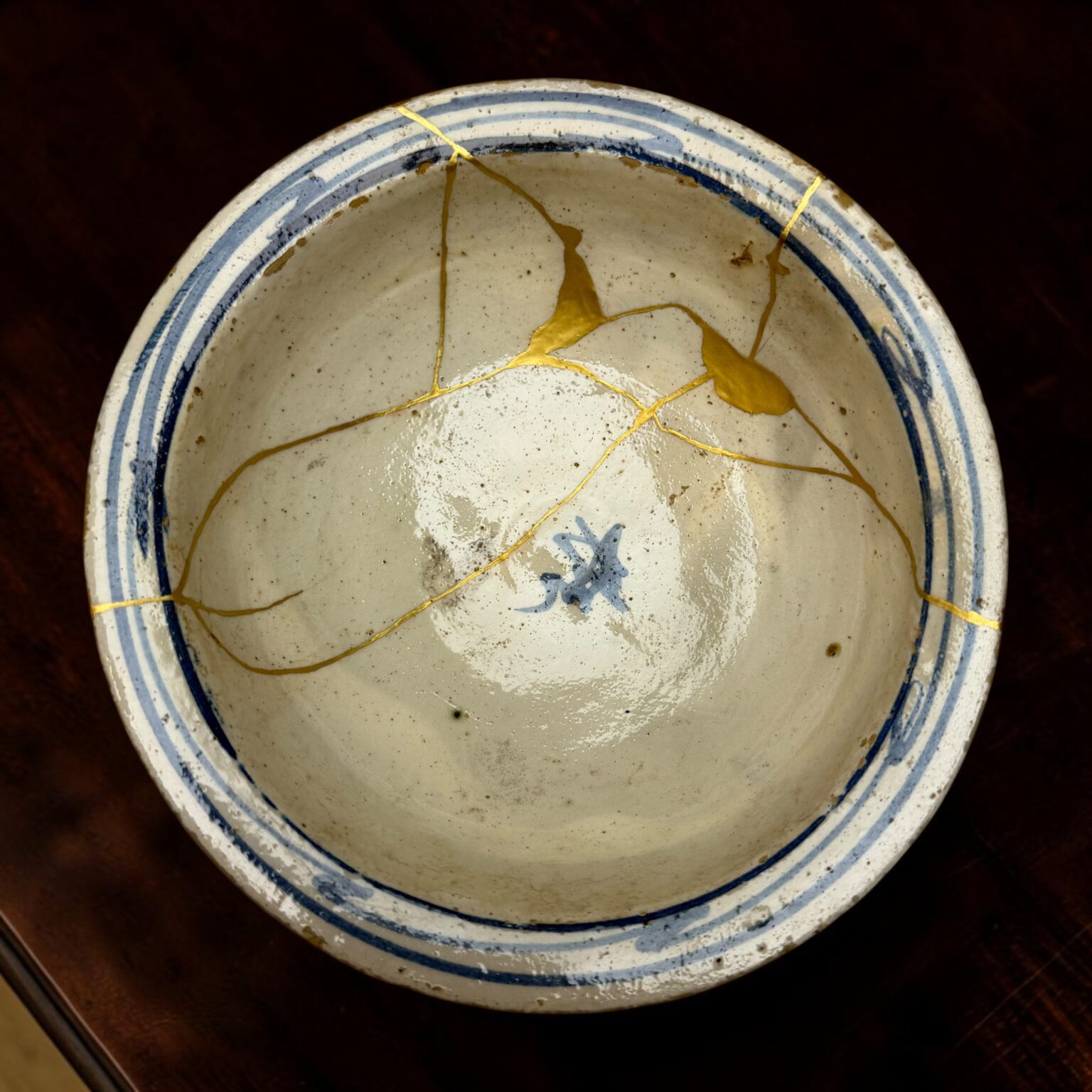 Close-up of a white ceramic plate with blue striped rim and gold repair seams (kintsugi) against a dark background, showing repaired cracks.",