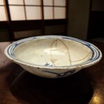 Close-up of a white ceramic bowl with blue decorative rim resting on a dark wooden table, background is blurred indoors.