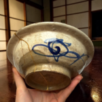 Close-up of a weathered ceramic bowl cradled in two hands, showing cracks and a blue decoration.