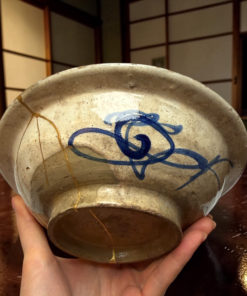 Close-up of a weathered ceramic bowl cradled in two hands, showing cracks and a blue decoration.