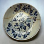 Blue-and-white ceramic plate with gold repair seams (kintsugi) and an intricate floral pattern, shown close-up against a light background.