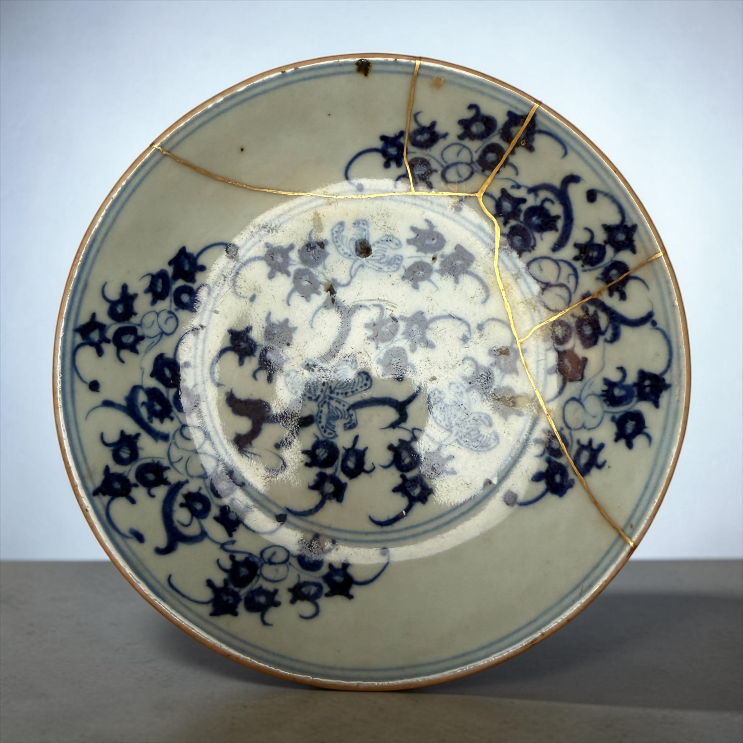 Close-up of a blue-and-white porcelain plate repaired with gold seams (kintsugi) on a gray surface.