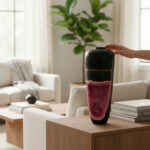 A tall black ceramic vase with pink-purple glaze at the bottom sits on a wooden table as a hand reaches to touch its rim in a modern living room.