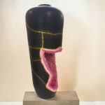 Black sculpture bust with gold vein cracks and a pink geode inset along the torso, displayed on a gray pedestal.