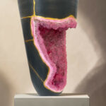 Close-up of a black ceramic vase cut open to reveal a pink crystal interior along a yellow seam, displayed on a pedestal.