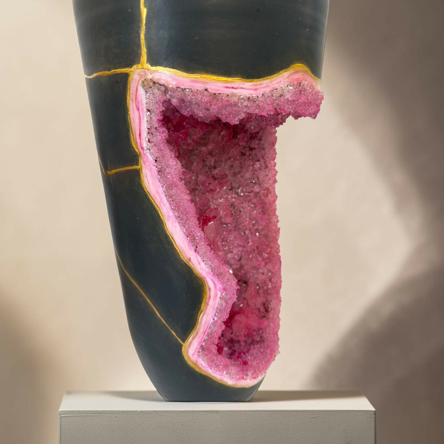 Close-up of a black ceramic vase cut open to reveal a pink crystal interior along a yellow seam, displayed on a pedestal.