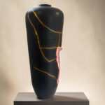 Black ceramic vase with gold vein cracks on a gray pedestal against a light background (art display)