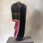 Black ceramic vase with pink crystalline growth along a yellow crack running vertically down its side, placed on a gray pedestal against a neutral wall.
