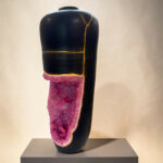 Black ceramic vase with yellow crackle glaze lines, sitting on a gray pedestal with pink crystalline material wrapped around its base, in a gallery setting.