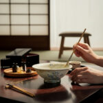 Hand using chopsticks to stir a ceramic bowl on a dark wooden table; a tray of small bottles sits nearby in a blurred background.