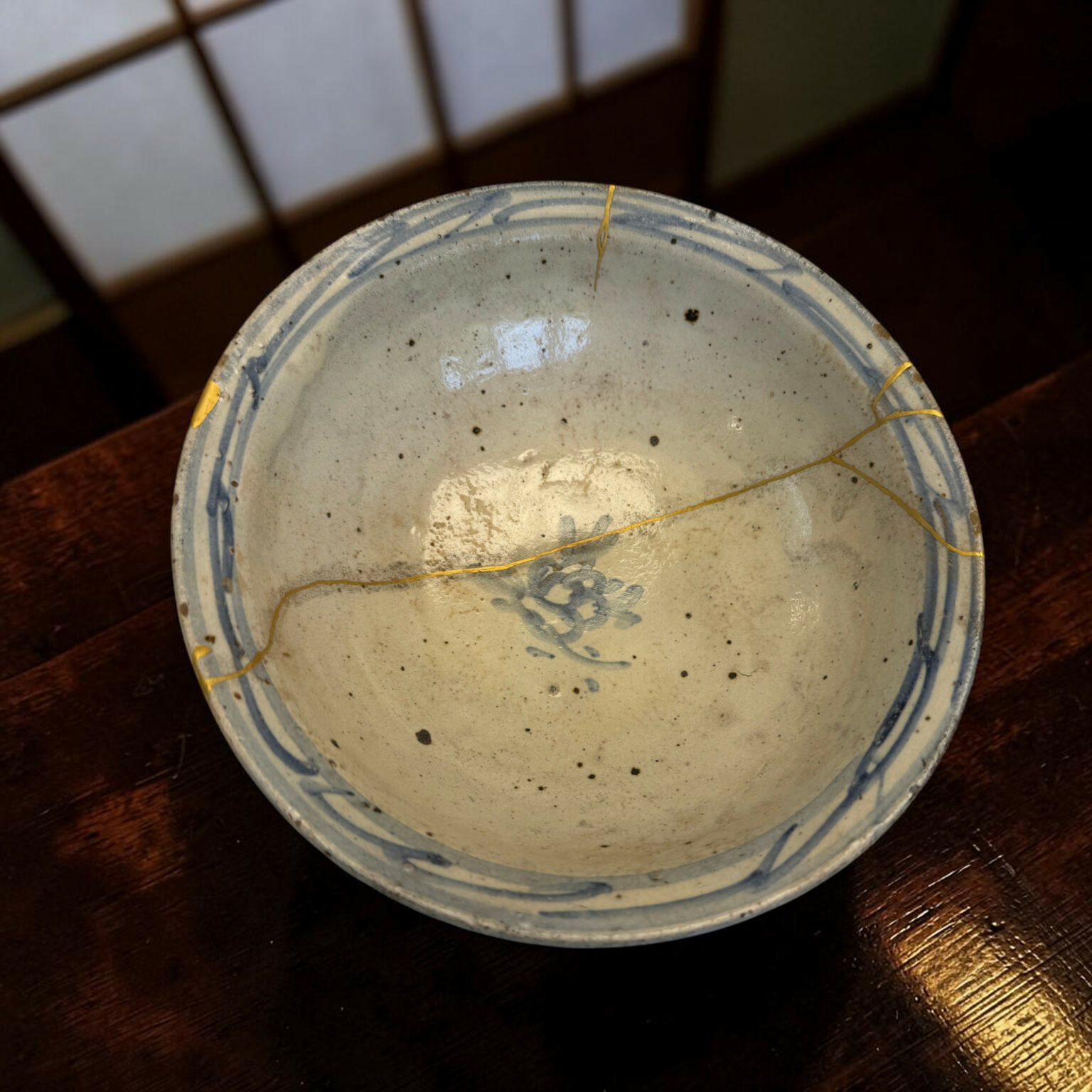 Close-up of a speckled ceramic plate with a blue rim on a dark wooden table, with a thin gold string along the edge several times.