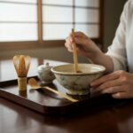 Person uses a wooden utensil to stir a ceramic bowl on a dark wooden tray; a bamboo whisk and small lidded pot sit nearby—traditional tea setup.