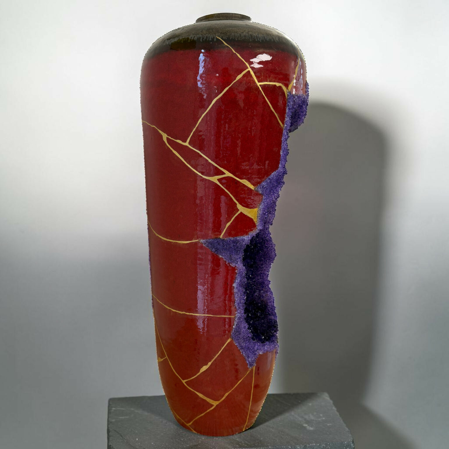Red ceramic vase with gold kintsugi cracks and a purple jagged edge along the side, displayed on a dark pedestal.