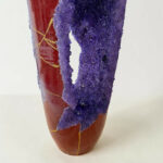 Close-up of purple crystalline material wrapped around a red ceramic vase with yellow string, on a pale background.