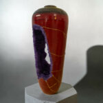 Close-up of a glossy red ceramic vase with purple crystalline formations along its side, placed on a pedestal.