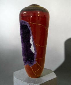 Close-up of a glossy red ceramic vase with purple crystalline formations along its side, placed on a pedestal.