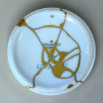 White porcelain plate repaired with gold seams (kintsugi) showing a network of gold cracks and a BERNAUD/BERNARDAUD mark visible in the glaze.