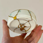 Person holds a cracked white porcelain plate with gold seams against a plain beige background
