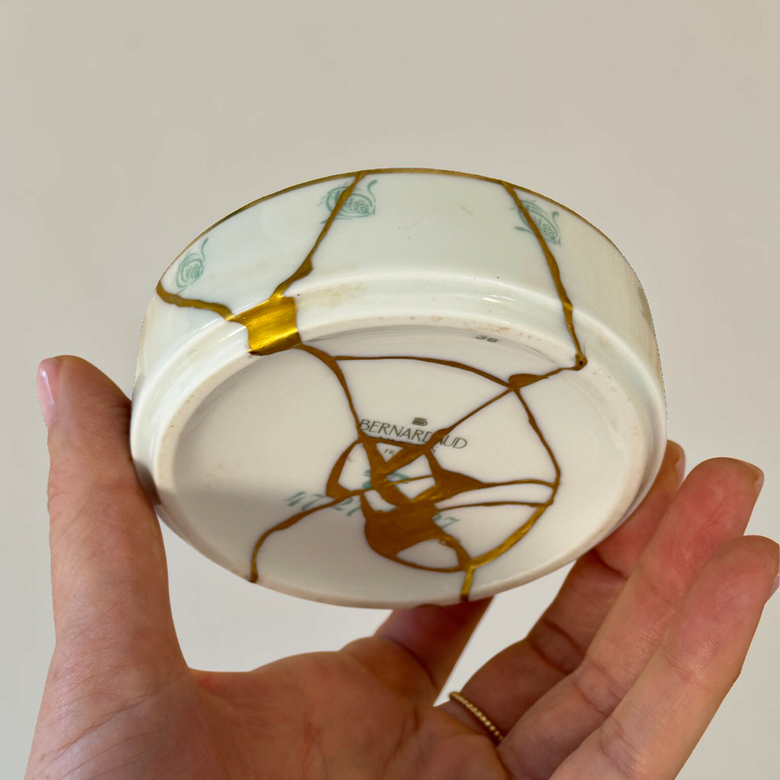 Person holds a cracked white porcelain plate with gold seams against a plain beige background